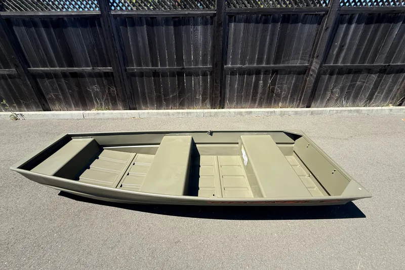 Slide: The Image of 2026 Tracker Grizzly 12 Jon boat on pavement, wooden fence background. - 2