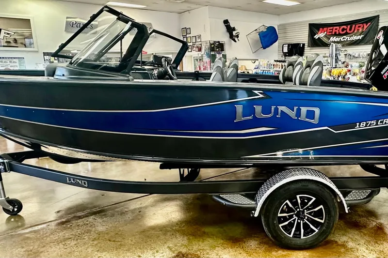 Slide: The Image of 2026 Lund 1875 Crossover XS boat in showroom, blue and black design, on trailer. - 2