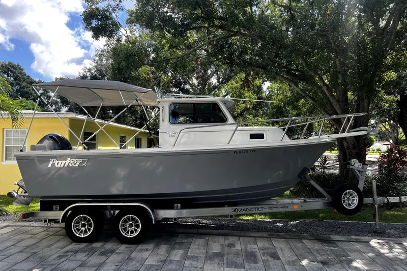 Slide: The Image of 2023 Parker 2120 Sport Cabin boat on trailer, parked outdoors under trees. - 1