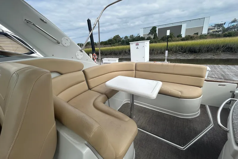 Slide: The Image of 2015 Crownline 350 SY boat interior with beige seating and table, docked by a grassy shoreline. - 14