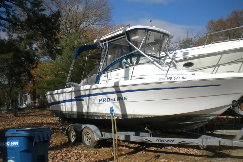 Slide: The Image of 1999 Pro-Line 24 Walkaround boat on trailer, parked outdoors with trees in the background. - 0
