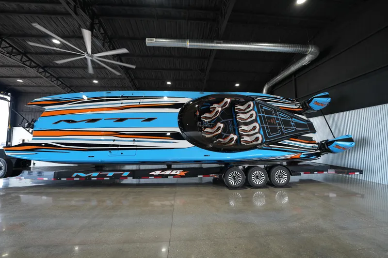 The Image of 2025 MTI 440X powerboat with vibrant blue and orange design on a trailer indoors. - 0