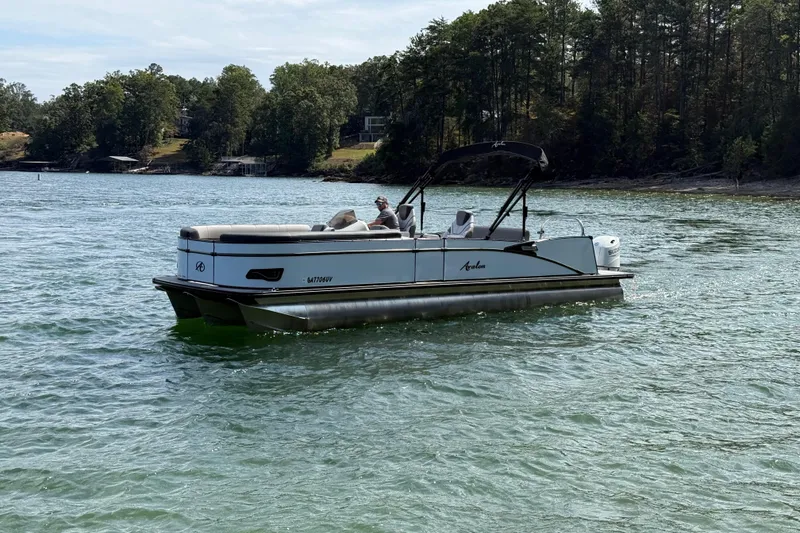 The Image of 2023 Avalon Catalina Versatile Rear Bench boat on a scenic lake with forested shoreline. - 2