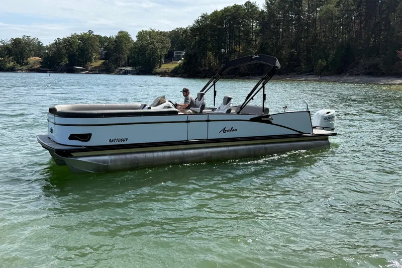 Slide: The Image of 2023 Avalon Catalina Versatile Rear Bench boat on a serene lake with lush green surroundings. - 4