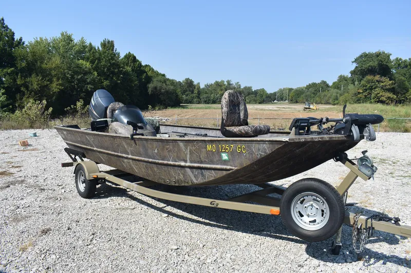 Slide: The Image of 2012 G3 1860 SC CAMO boat on trailer, parked on gravel with trees in background. - 3