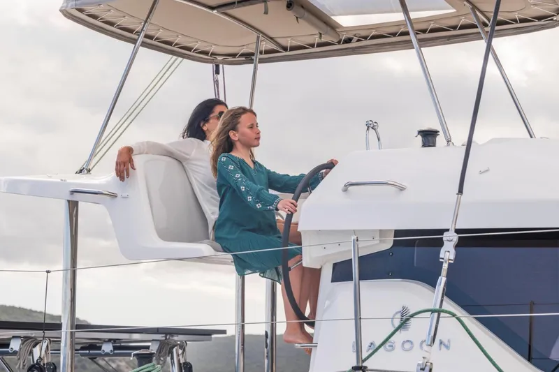 Slide: The Image of Young girl steering 2026 Lagoon 38 catamaran, with adult seated nearby, under cloudy sky. - 5