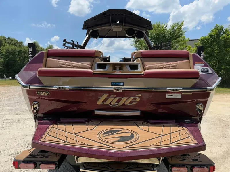 Slide: The Image of 2024 Tigé Ultré 25ZX boat, rear view, maroon and tan, parked outdoors under blue sky. - 6