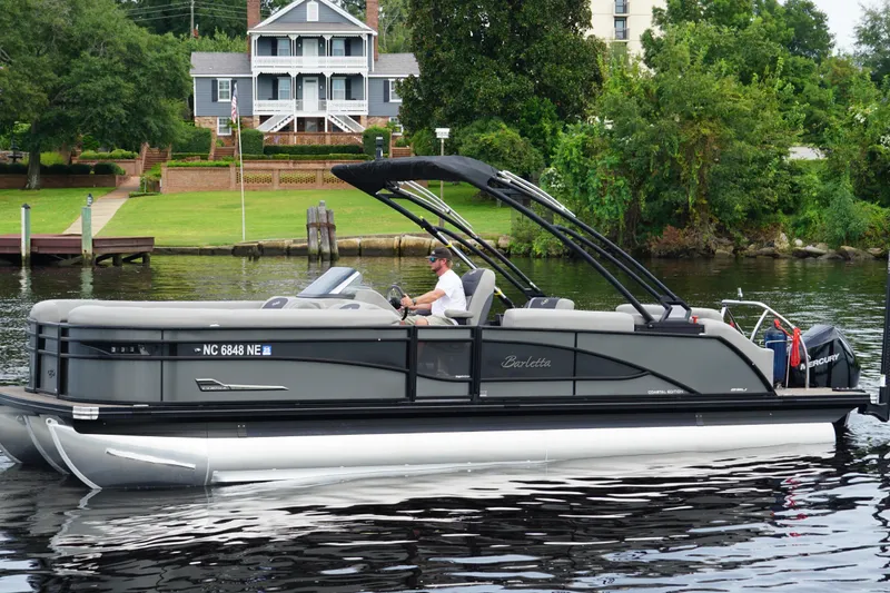 The Image of 2023 Barletta Corsa 25U pontoon boat cruising on a calm river near a scenic shoreline. - 1