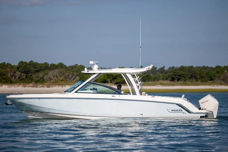 Slide: The Image of 2023 Boston Whaler 320 Vantage boat cruising on calm waters near a forested shoreline. - 10
