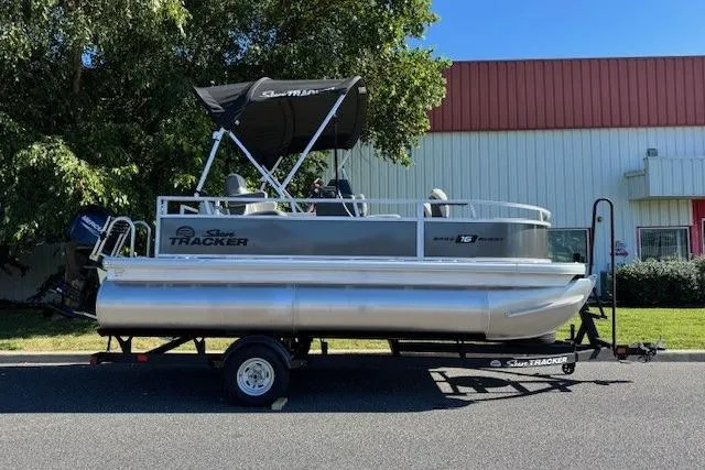 The Image of 2026 Sun Tracker Bass Buggy 16 XL Select pontoon boat on trailer, parked outdoors. - 0