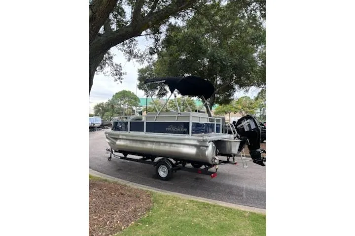 Slide: The Image of 2026 Sun Tracker Bass Buggy 16 XL Select pontoon boat on trailer, parked under trees. - 2