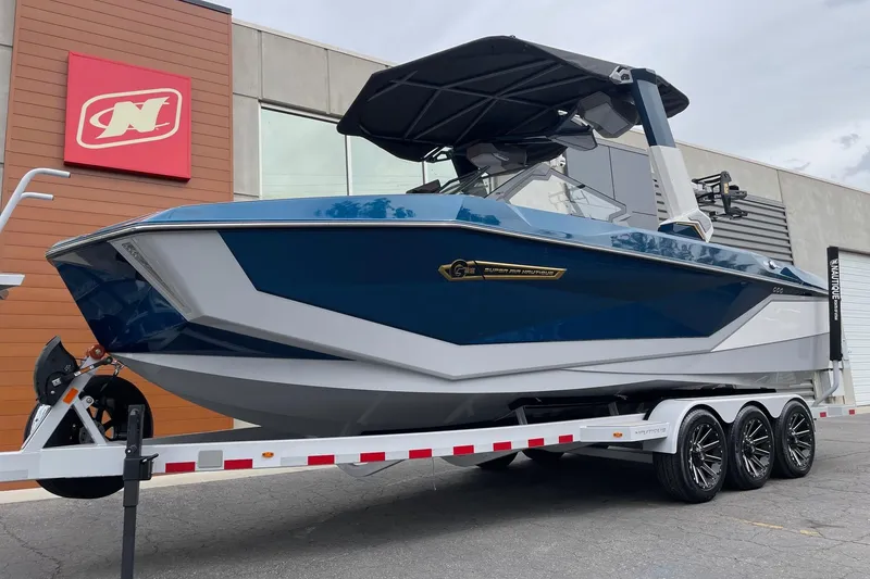 Slide: The Image of 2026 Nautique Super Air Nautique G25 boat on trailer, blue and white design, parked outside. - 2