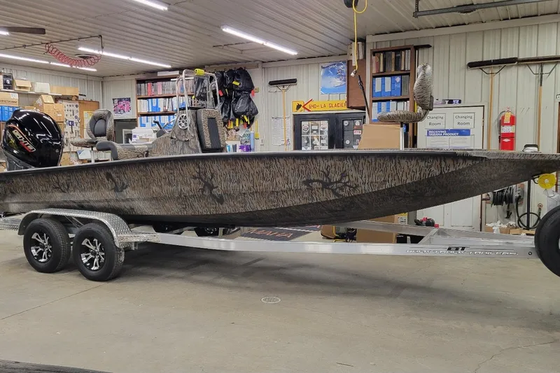 The Image of 2026 Xpress H22B Bay boat in a garage, featuring a camouflage design and trailer. - 1
