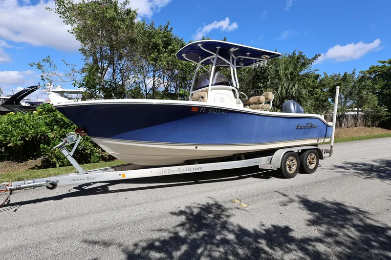 The Image of 2019 NauticStar 2302 Legacy boat on trailer, parked on roadside under blue sky. - 0