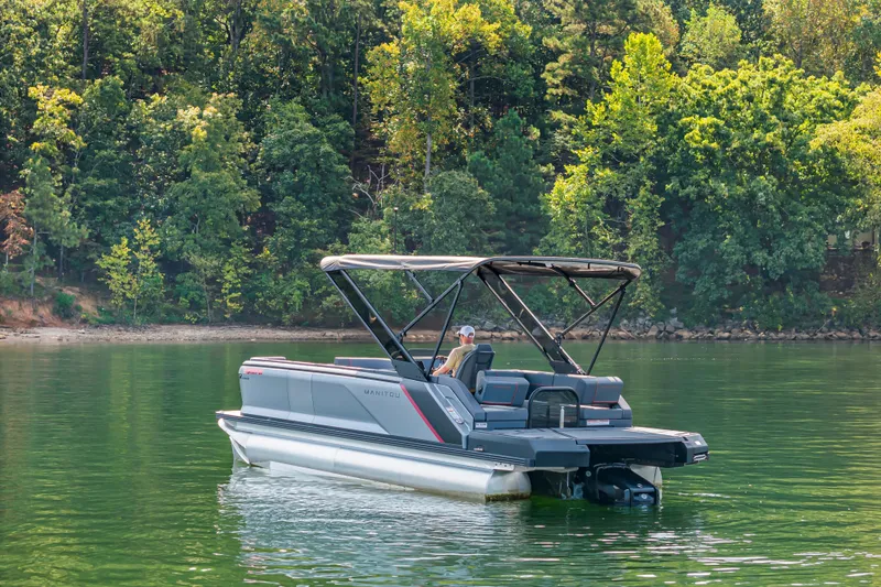 Slide: The Image of 2024 Manitou 22 Explore Max Bench pontoon boat on a serene lake with lush forest backdrop. - 3