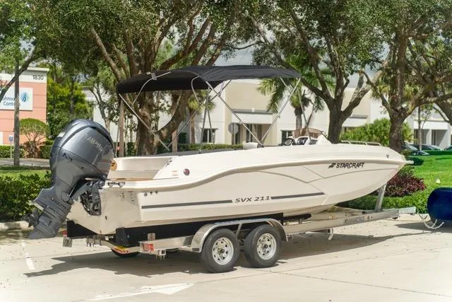 Slide: The Image of 2026 Starcraft SVX 211 OB boat on trailer, parked outdoors with trees in background. - 6
