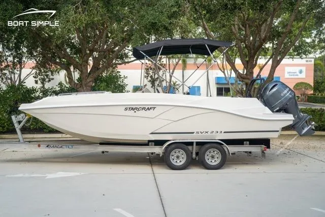 Slide: The Image of 2023 Starcraft SVX 231 OB DH boat on trailer, parked outdoors with trees in background. - 0
