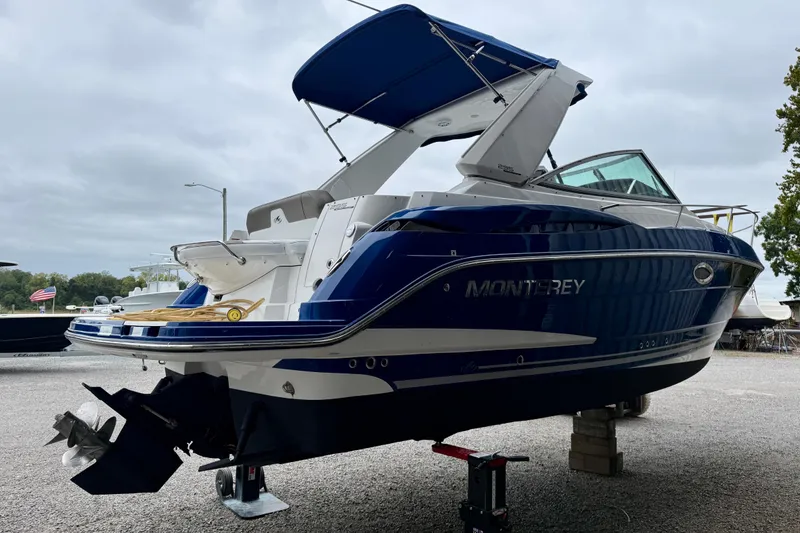 Slide: The Image of 2019 Monterey 295 Sport Yacht in blue, docked on a gravel surface under cloudy skies. - 5