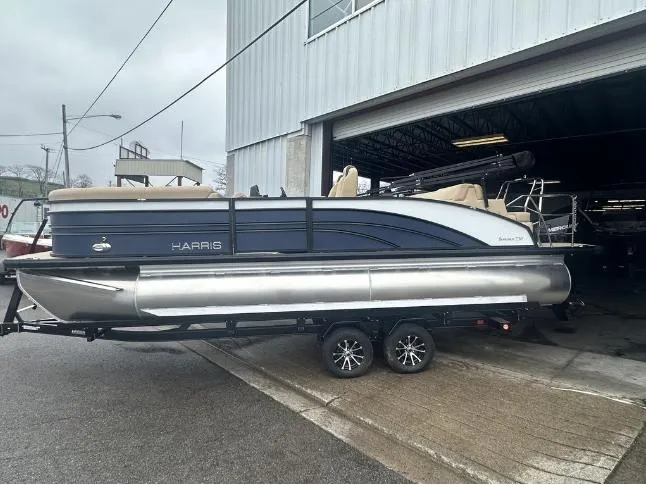 Slide: The Image of 2024 Harris Sunliner 230 pontoon boat on a trailer outside a warehouse. - 1