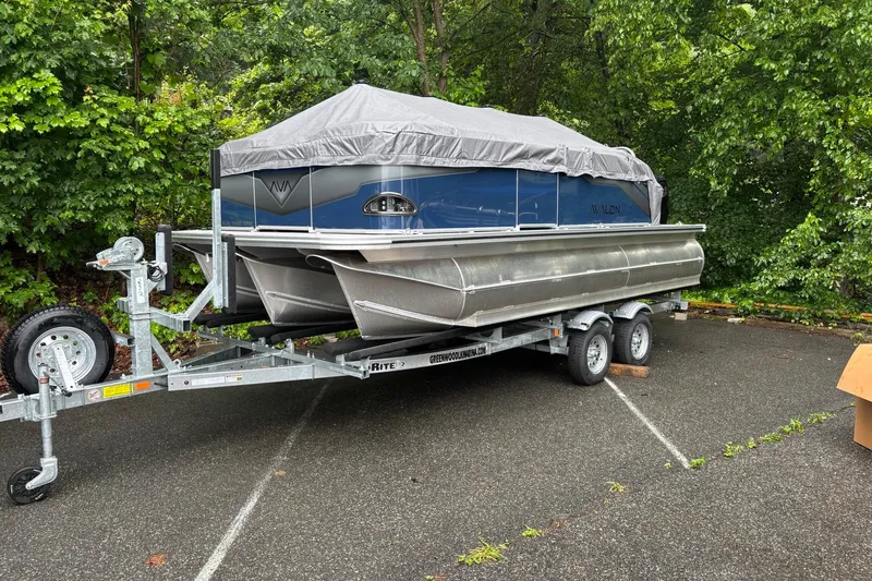 Slide: The Image of 2025 Avalon Venture Cruise Rear Bench pontoon boat on trailer, covered, in wooded parking area. - 1