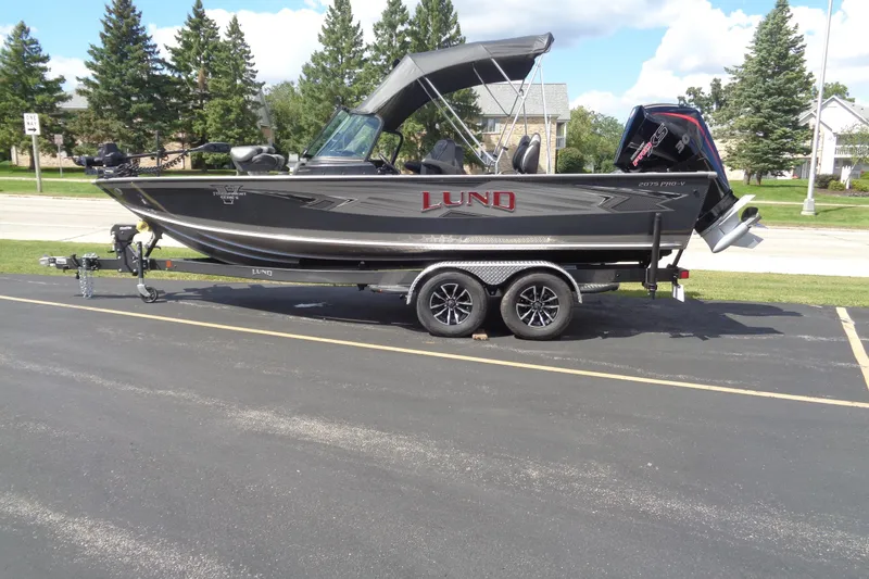 The Image of 2026 Lund 2075 Pro-V Sport boat on trailer, parked outdoors, with trees in background. - 1