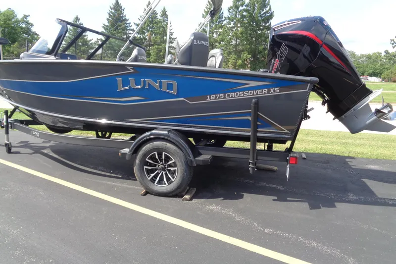Slide: The Image of 2026 Lund 1875 Crossover XS boat on trailer, blue and black design, parked outdoors. - 2