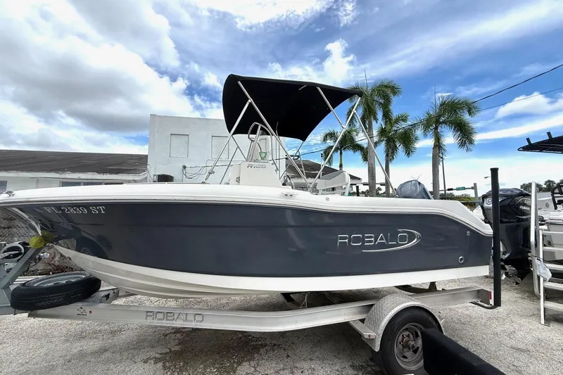 The Image of 2020 Robalo R180 Center Console boat on trailer, under cloudy sky with palm trees. - 0
