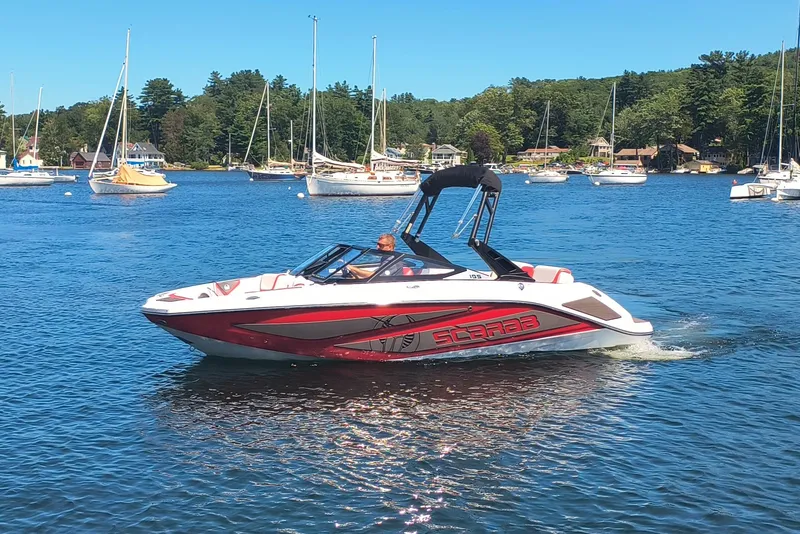 Slide: The Image of 2023 Scarab 195 ID boat cruising on a scenic lake with sailboats in the background. - 2