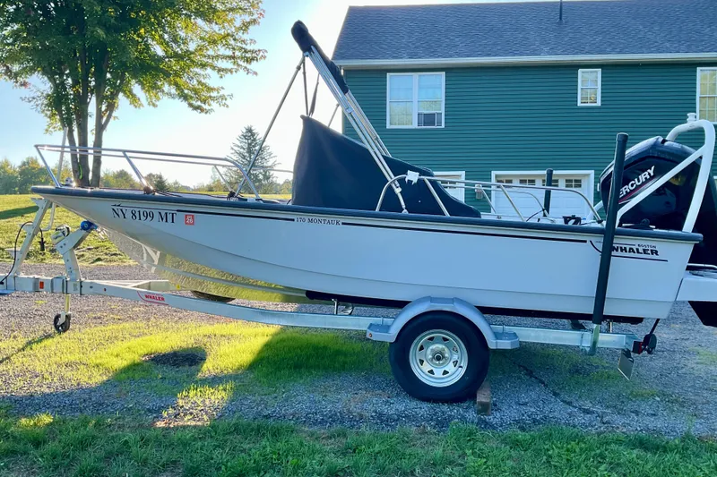 Slide: The Image of 2024 Boston Whaler 170 Montauk boat on trailer, parked in front of a green house. - 20