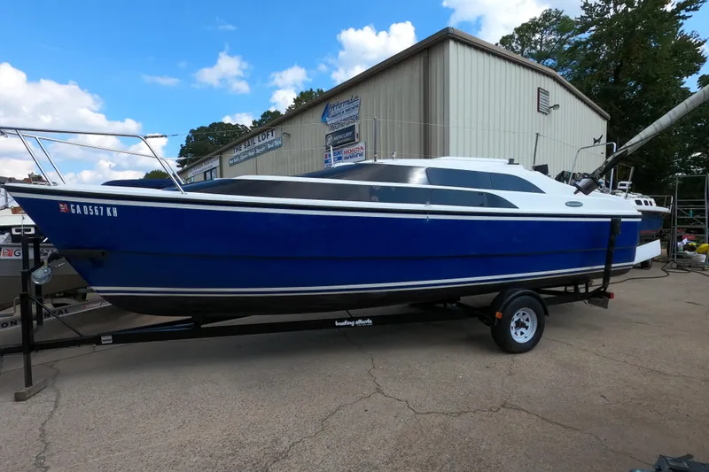 The Image of Blue 2007 MacGregor 26M sailboat on trailer, parked outside a building under a clear sky. - 0