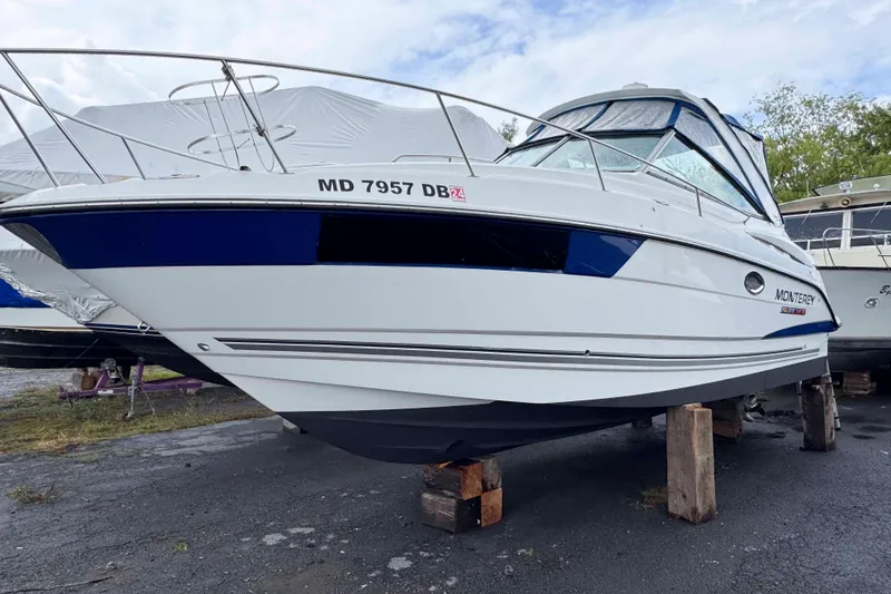 The Image of 2019 Monterey 295 Sport Yacht on blocks, white hull, blue accents, parked outdoors. - 0