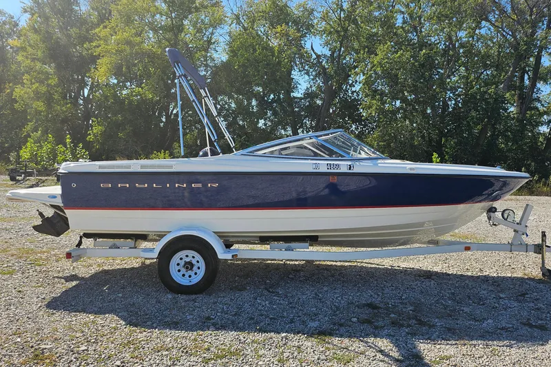 Slide: The Image of 2004 Bayliner 215 Bowrider on trailer, parked outdoors with trees in background. - 3