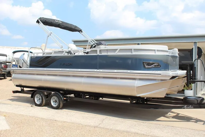 Slide: The Image of 2026 Avalon LSZ Cruiser Rear Bench 23 pontoon boat on trailer, parked outdoors. - 7
