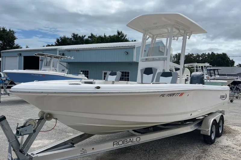 Slide: The Image of 2024 Robalo 226 Cayman boat on trailer, parked outdoors near a blue building. - 2