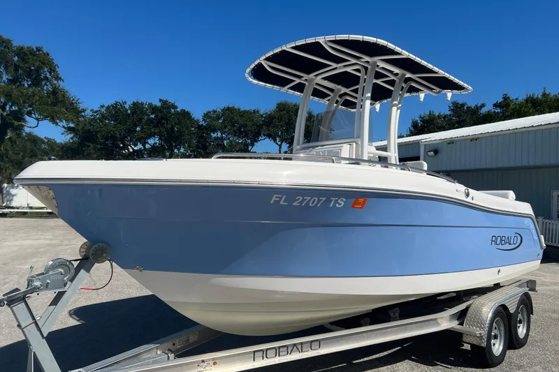 The Image of 2024 Robalo R222 Center Console boat on trailer, blue and white design, sunny day. - 0
