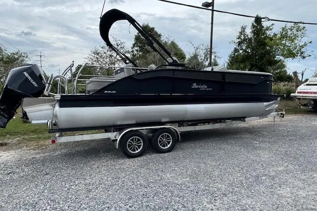 Slide: The Image of 2026 Barletta ARIA 24 UC pontoon boat on a trailer, parked outdoors. - 2