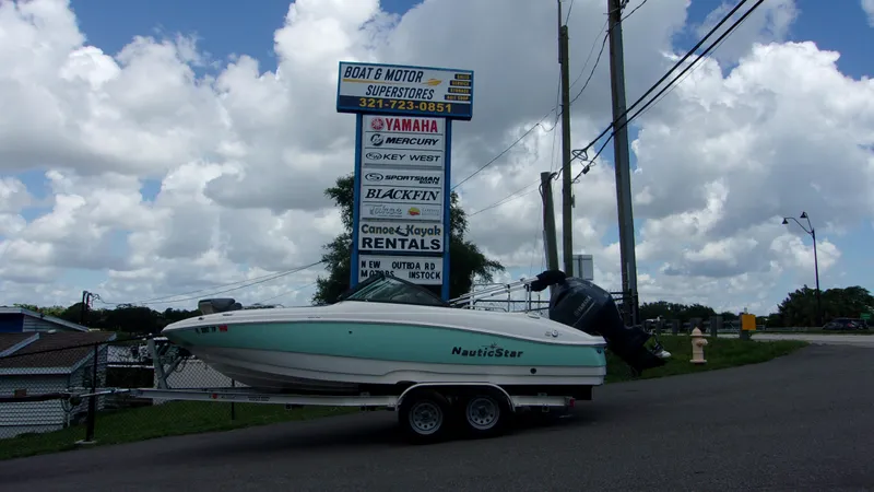 The Image of 2023 NauticStar 203 DC boat on trailer near boat dealership sign under cloudy sky. - 0