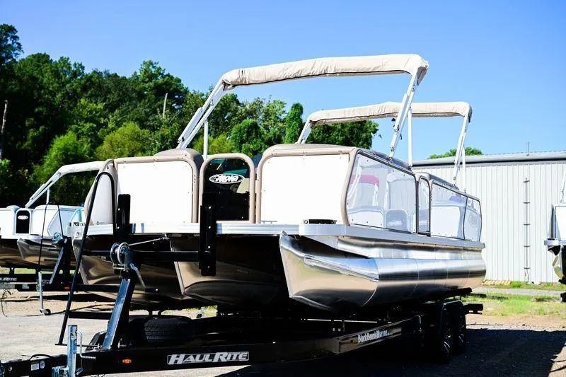 Slide: The Image of 2023 Aloha Pontoon Mahalo 23 on trailer, parked outdoors, with trees and building in background. - 2