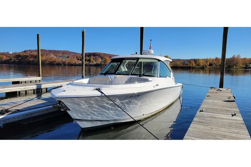 Slide: The Image of 2024 Pursuit DC 326 Dual Console boat docked on a serene lake with autumn foliage. - 2
