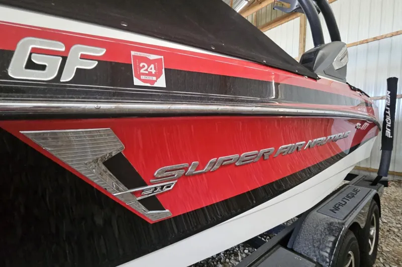 Slide: The Image of 2016 Nautique Super Air Nautique 210 boat, red and black design, parked indoors. - 11