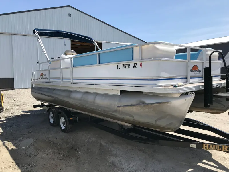 The Image of 2006 Sunset Bay Cruise Deluxe pontoon boat on trailer near a large building. - 1