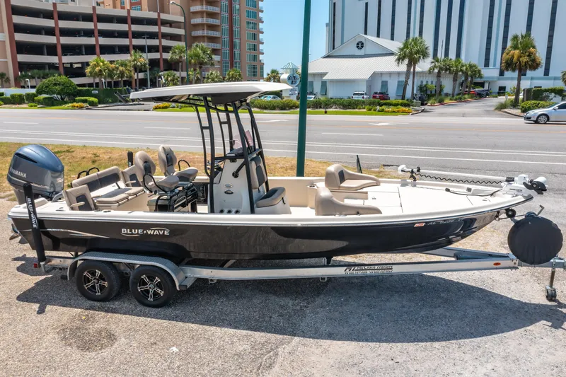 Slide: The Image of 2020 Blue Wave 2400 PureBay boat on trailer, parked near buildings and road. - 0