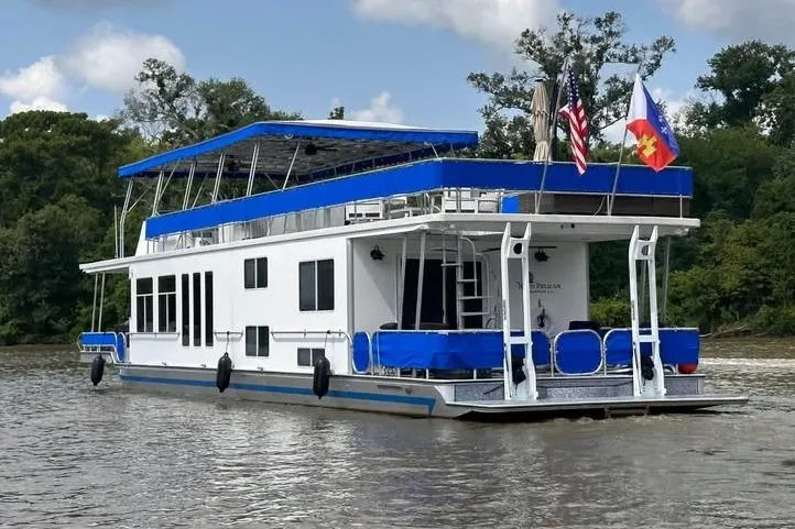 The Image of 1999 Sharpe houseboat on a river, featuring blue accents and flags, surrounded by trees. - 0
