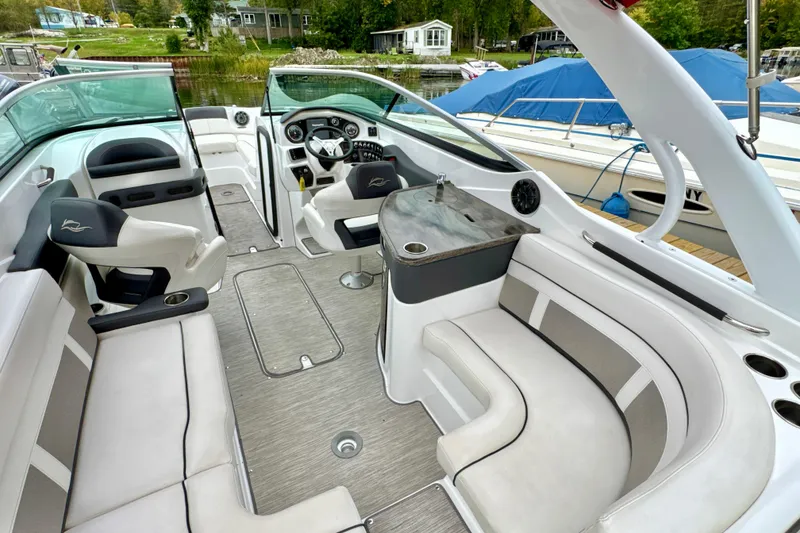 Slide: The Image of 2018 Rinker 29QX BR boat interior with seating, steering wheel, and table, docked by a lake. - 7