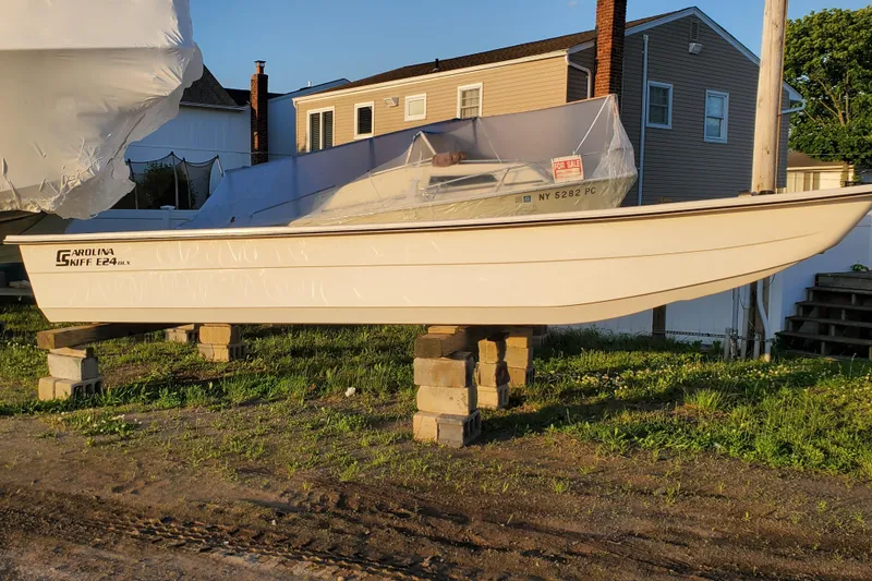 Slide: The Image of 2024 Carolina Skiff E24DLX boat on blocks, outdoor setting, for sale sign visible. - 1