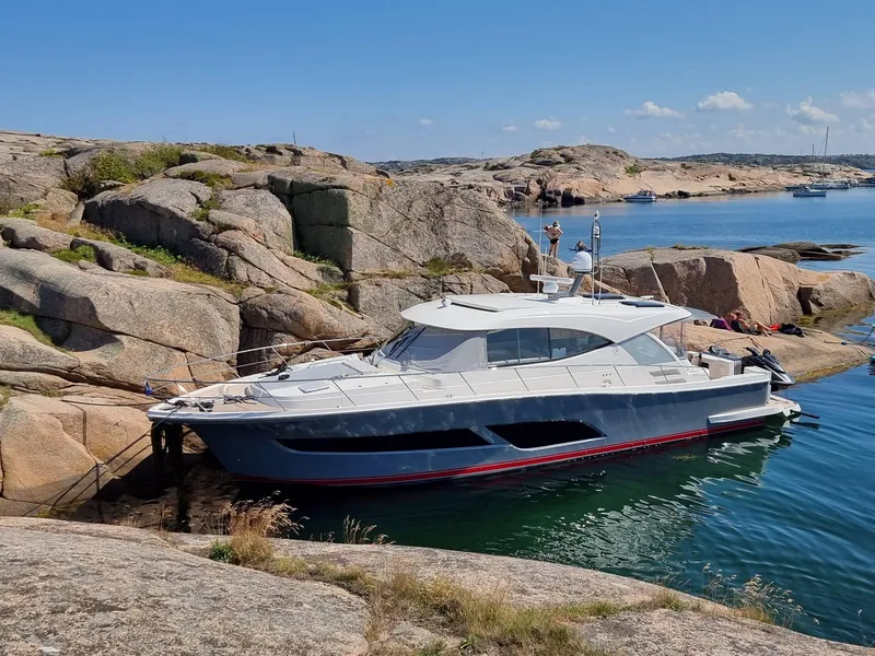 Slide: The Image of 2022 Riviera 505 SUV yacht docked by rocky shoreline under clear blue sky. - 0