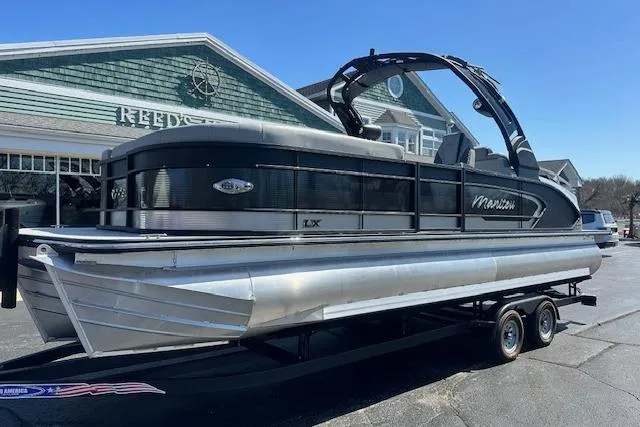The Image of 2024 Manitou LX 25 SRS pontoon boat on trailer, parked outside a building. - 0