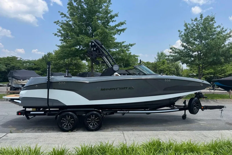 The Image of 2025 MasterCraft NXT24 boat on trailer, parked outdoors, surrounded by trees and clear sky. - 0