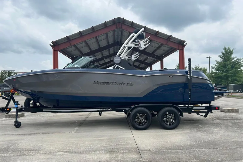 The Image of 2025 MasterCraft NXT23 boat on trailer under a metal canopy, cloudy sky background. - 0