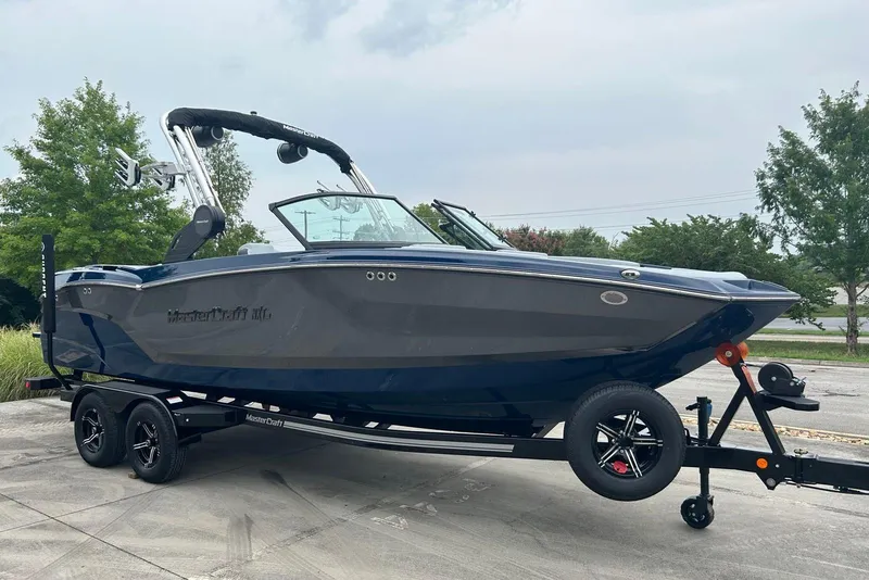 Slide: The Image of 2025 MasterCraft NXT23 boat on trailer, parked outdoors with trees in background. - 1